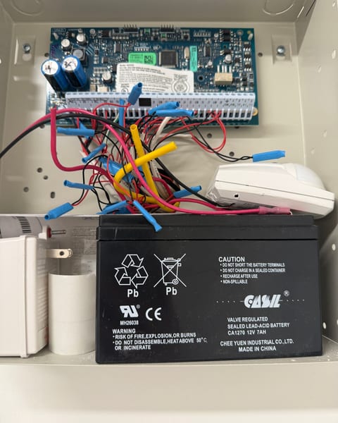 An open alarm panel enclosure: the control board with its wiring terminals at the top and a 12-volt backup battery below.