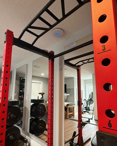 A white UniFi access point mounted flush on the textured ceiling of a home gym, its blue status ring lit, above a red power rack with a black pull-up bar and wall mirrors.