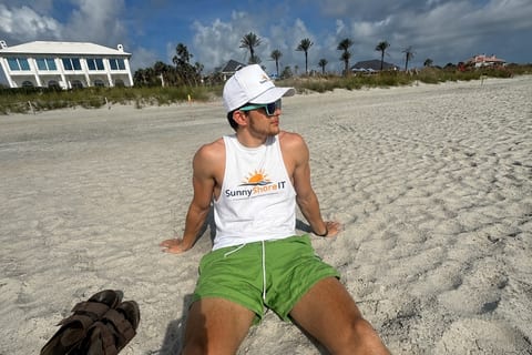 Logan sitting on the sand at the beach in a white SunnyShoresIT tank top, a white cap and sunglasses, with dune grass, palm trees and beach houses behind him.