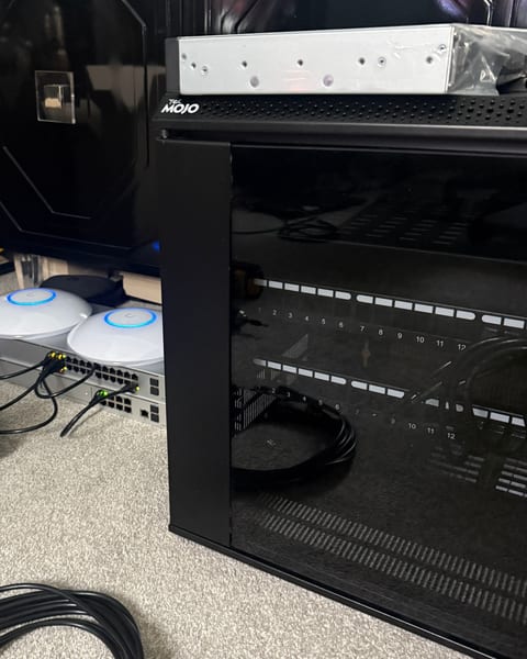 A small black network rack with a glass door and two numbered patch panels, next to two ceiling access points and a switch on the carpet, and a coil of black patch cables in front.