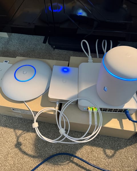 UniFi equipment set out on its boxes with status lights on: a round access point, a wall-plate access point, a gateway and a switch, cabled with short white patch cords.